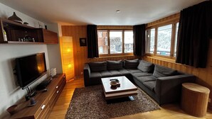 Ein modernes Wohnzimmer mit einer Eckcouch, einem Flachbildfernseher, einem Holztisch und großen Fenstern mit Vorhängen.