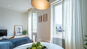 Een moderne woonkamer met een witte bank, een eettafel met een schaal groene appels, een flatscreen-tv en een groot raam met transparante gordijnen.