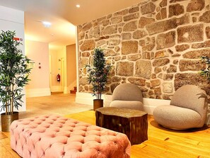 Una hall con una parete in pietra, due piante in vaso, un pouf trapuntato e un tavolino rotondo in legno.