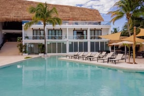 Un área de piscina con sillas de descanso, un edificio con ventanas amplias y una estructura de techo de paja.