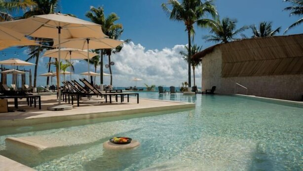 A clear pool with lounge chairs and umbrellas, surrounded by palm trees and a thatched-roof structure. - The Beachfront by The Fives Hotels Á la Carte - Adults Only (Playa del Carmen)