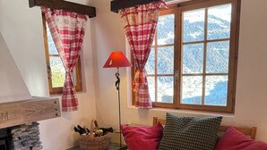 A cozy living room with a stone fireplace, a wooden sofa with red cushions, a coffee table with cups and a jar of cookies, and a view of snow-covered mountains through a window with red curtains. - Comfortable chalet for 5 persons : Living room with fireplace - kitchen with dishwasher, microwave a (Grimentz)