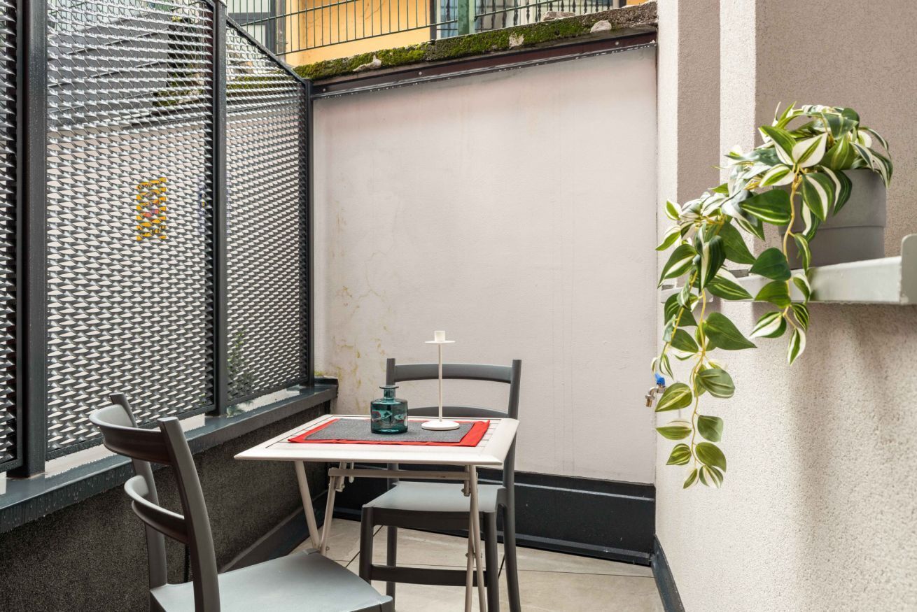 Een klein terras buiten met een tafel en twee stoelen, een metalen hekwerk en een pot met een plant.