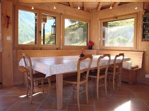 A wooden dining area with a long table and chairs, large windows with a view of greenery, and a bench with a vase of flowers. - Chalet high standing close to the slopes for 12 persons : 1. floor : living-room with wood stove - k (Grimentz)