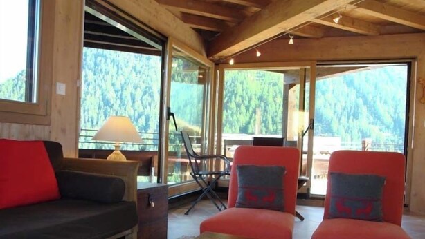 Ein gemütliches Zimmer mit Holzbalkendecke, einem großen Fenster mit Blick auf einen Wald, einer Couch, zwei Sesseln und einem Tisch.