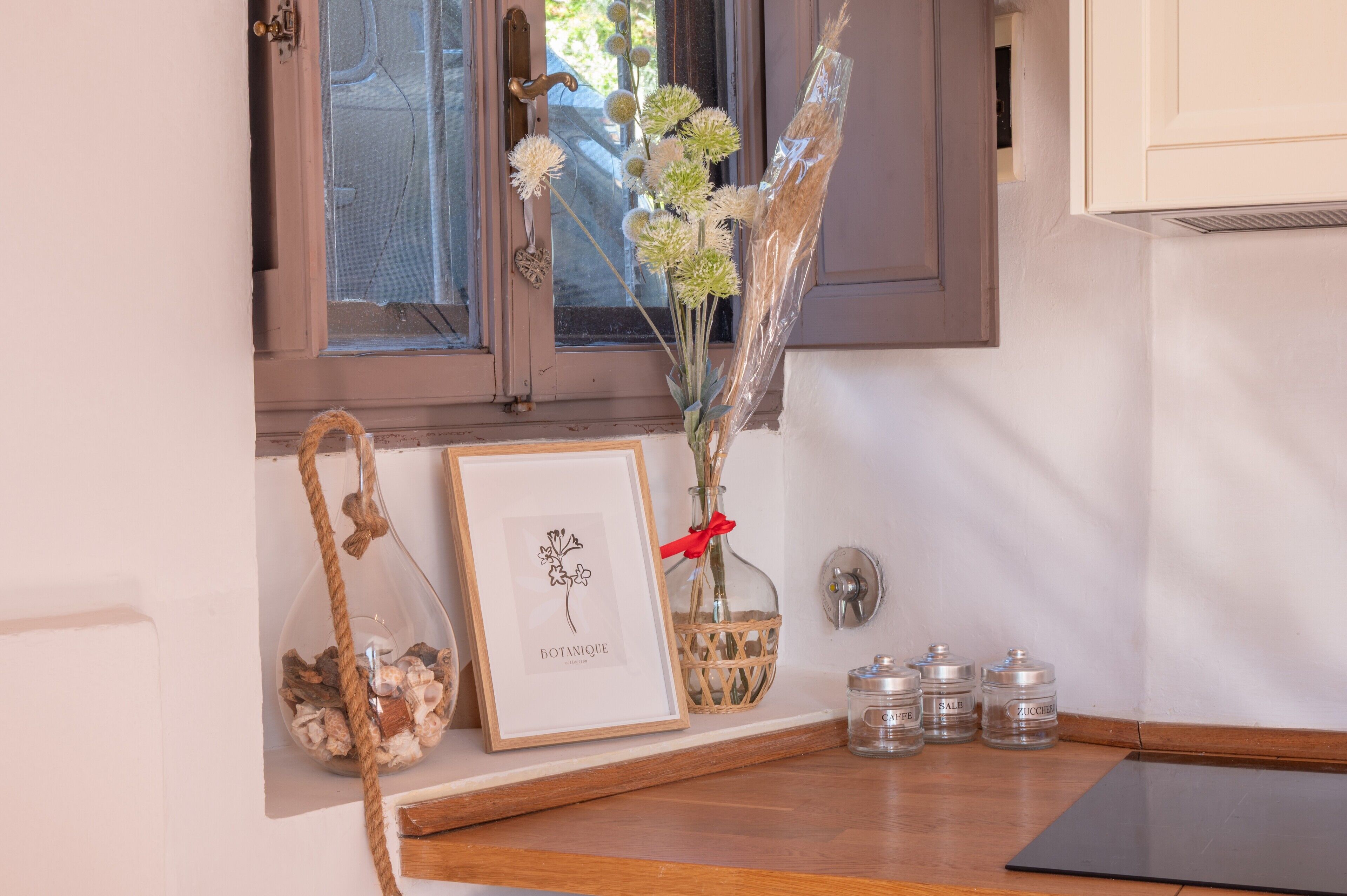 A framed botanical print, a vase with dried flowers, a wicker basket, and three glass jars labeled 'CANDLE,' 'SALE,' and 'ZUCCHERO' on a wooden surface near a window.