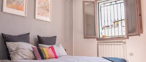 A bedroom with a bed, two framed artworks, a window with an open shutter, and a radiator.