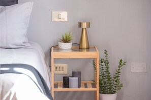 Ein ordentlich bezogenes Bett mit weißem Kopfkissen, einer kleinen Topfpflanze, einer Lampe und einem Regal mit Kerzen und einer Pflanze.