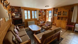 Ein gemütliches Blockhäuschen-Interieur mit einer Couch, Sesseln, einem Esstisch, einem Fernseher und einem Bücherregal.
