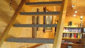 Interior acogedor de una cabaña con cama en altillo, escalera de madera y estantería llena de libros.