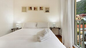A hotel room with a bed, two bedside tables, a wall-mounted air conditioner, and a window with a view of buildings and a mountain. - Apartment 'Machico Beach Front' with Sea View, Wi-Fi and Air Conditioning (Machico)