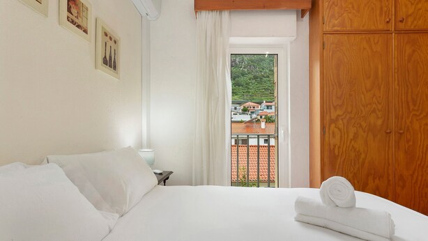 A hotel room with a bed, a nightstand, a window with a view of buildings and a mountain, and wooden furniture. - Apartment 'Machico Beach Front' with Sea View, Wi-Fi and Air Conditioning (Machico)