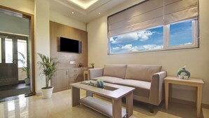 A modern living room with a sofa, coffee table, and a TV mounted on the wall. There is a window with a view of the sky and clouds.