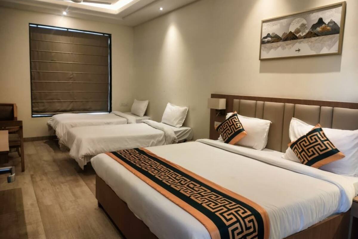 Habitación de hotel con tres camas, ventilador de techo, ventana grande con persianas y un cuadro de un paisaje montañoso en la pared.