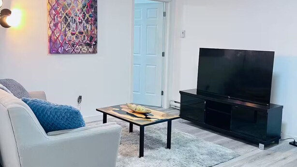 Ett vardagsrum med en vit soffa, ett litet soffbord, en platt-TV och en färgstark, abstrakt tavla på väggen.