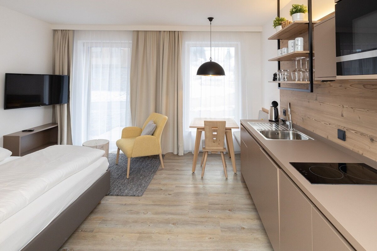 A modern hotel room with a bed, a TV, a dining table, a chair, and a kitchen area with a sink and a stove.