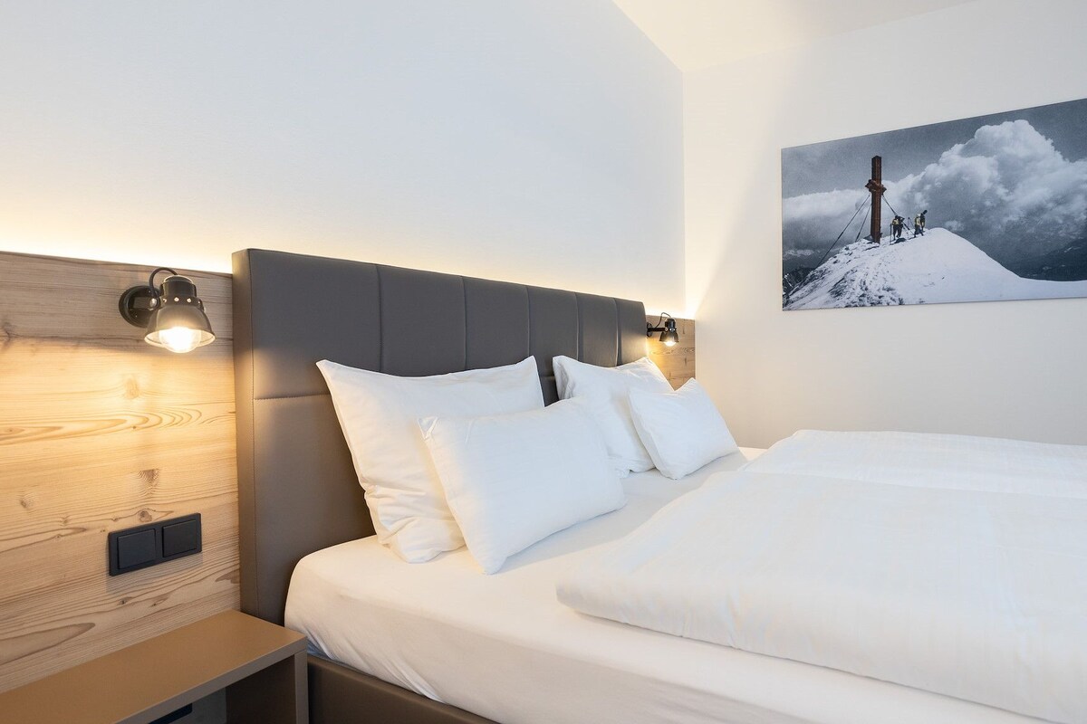 A hotel room with a bed, a nightstand, a wall-mounted lamp, and a framed picture of a mountain.