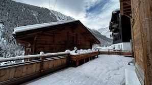 Eine Holzhütte mit Balkon, umgeben von schneebedeckten Bergen und einer Seilbahn im Hintergrund.