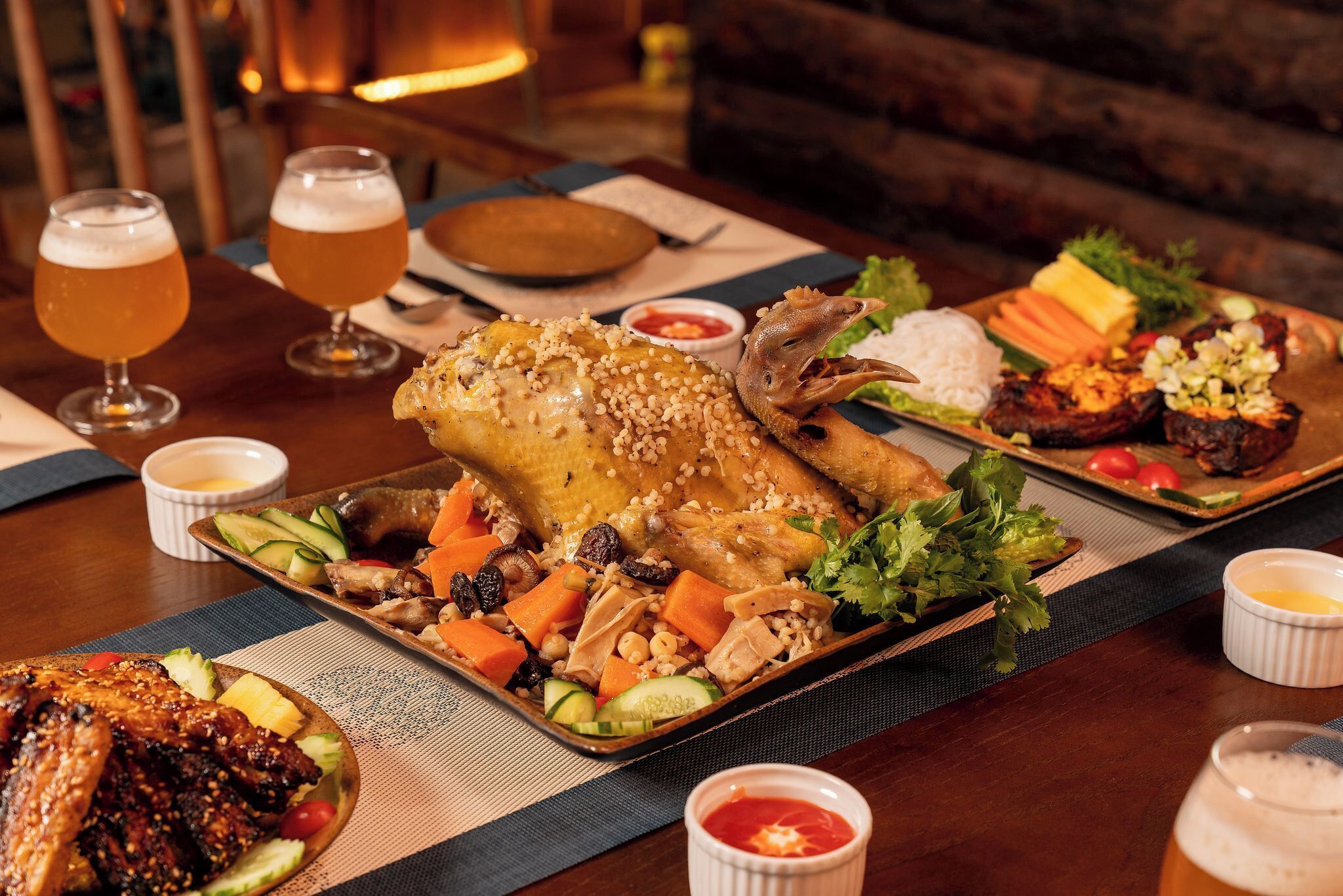 A dining table with various dishes, including roasted chicken, vegetables, and a whole fish, along with two glasses of beer.
