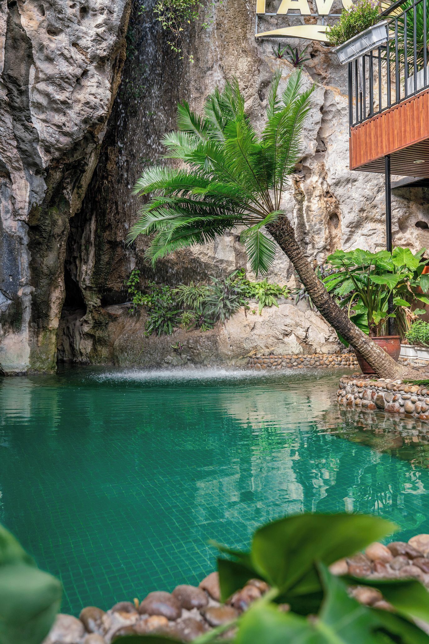 Una piscina amb aigua cristal·lina, envoltada de roques i vegetació, amb un penya-segada rocosa i un balcó amb barana de fusta.