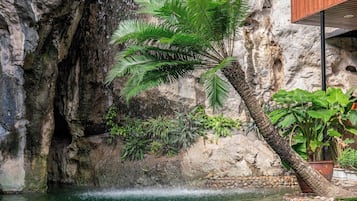 Una piscina con agua cristalina, rodeada de rocas y plantas, con un risco rocoso y un balcón con barandilla de madera.