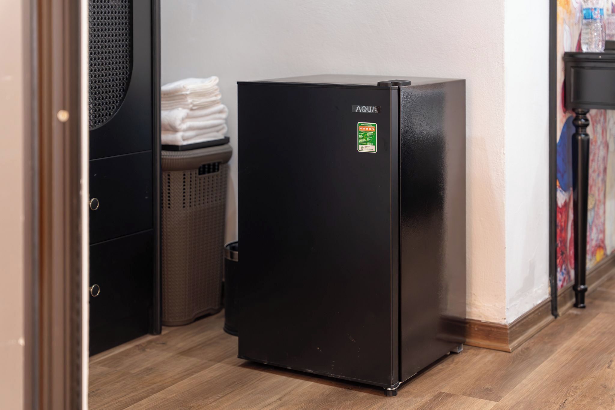 A black refrigerator in a room with wooden flooring, a trash bin, and a wall with a colorful painting.