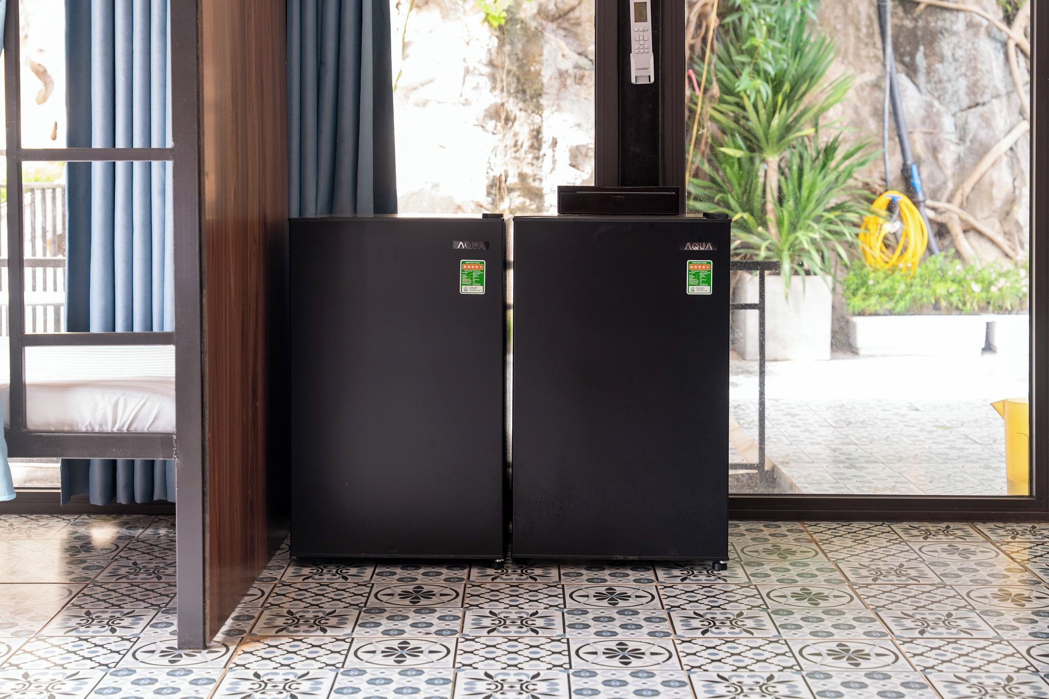 A room with two black refrigerators, patterned floor tiles, a window with curtains, and a view of a garden.