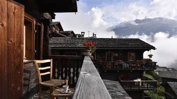Un balcon en bois avec une chaise et une table, donnant sur un village aux maisons traditionnelles, avec une montagne en arrière-plan.