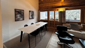 Eine Holzhütte mit Esstisch, Stühlen, einer Couch und großen Fenstern mit Blick auf Bäume.