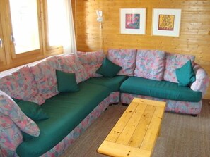 Ein gemütliches Zimmer mit einem grünen Ecksofa, einem Holztisch und zwei gerahmten Kunstwerken an der Holzwand.