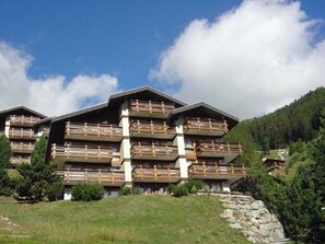 A multi-story hotel with wooden balconies and a stone foundation, surrounded by greenery and a mountainous backdrop. - Comfortable 3 room apartment for 5 persons : Living room with fireplace - kitchen with dishwasher, c (Grimentz)