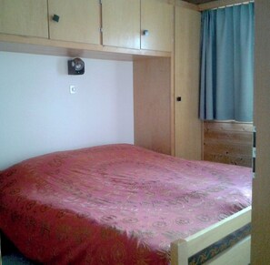 Ein Schlafzimmer mit einem Bett, Holzschränken, einem kleinen Fenster mit Vorhängen und einer Wandleuchte.