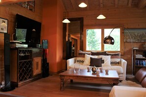 Ein gemütliches Wohnzimmer mit Holzboden, einem weißen Sofa mit gemusterten Kissen, einem Holztisch und einem Fernseher auf einer Holzkommode.