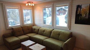 Ein Wohnzimmer mit einer Eckcouch, zwei Holztischen und großen Fenstern mit Blick auf ein Haus und Bäume.