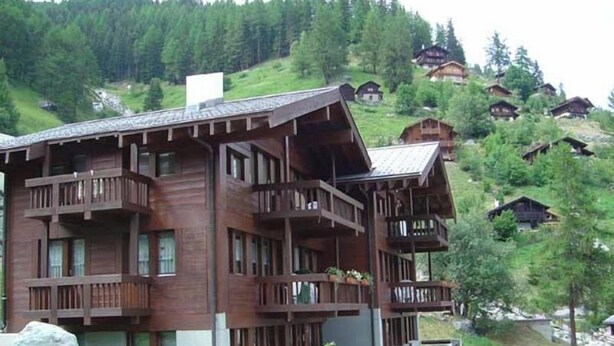 Eine mehrstöckige Holzhütte mit Balkonen, eingebettet in eine Berglandschaft, im Hintergrund sind weitere ähnliche Chalets zu sehen.