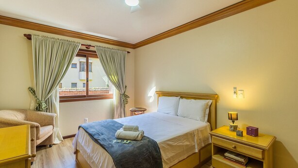 Una camera d'albergo con un letto, un comodino, una sedia, una finestra con tende e una ventola a soffitto.