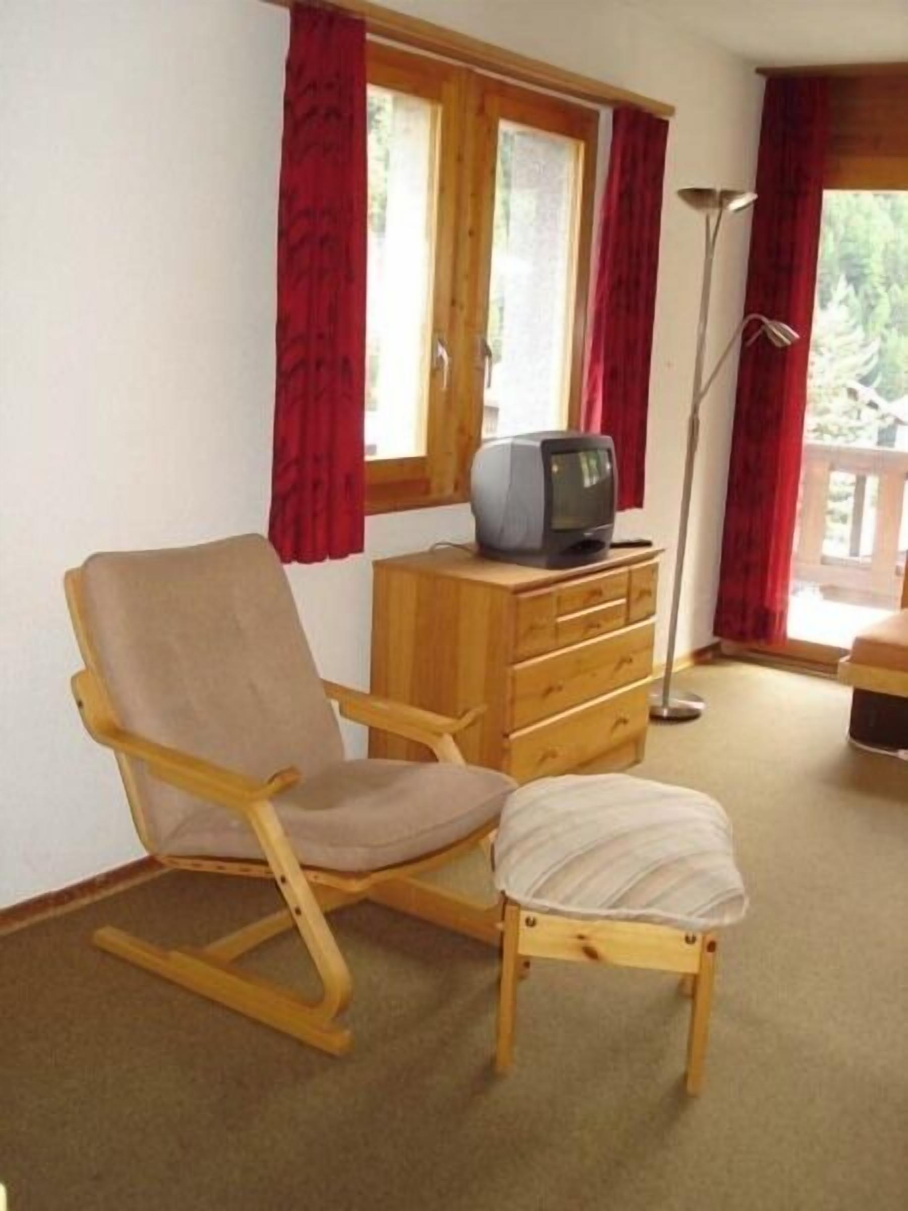 Una habitación con una mecedora de madera, un mueble bajo de madera con un televisor encima, una lámpara de pie y una ventana con cortinas rojas.