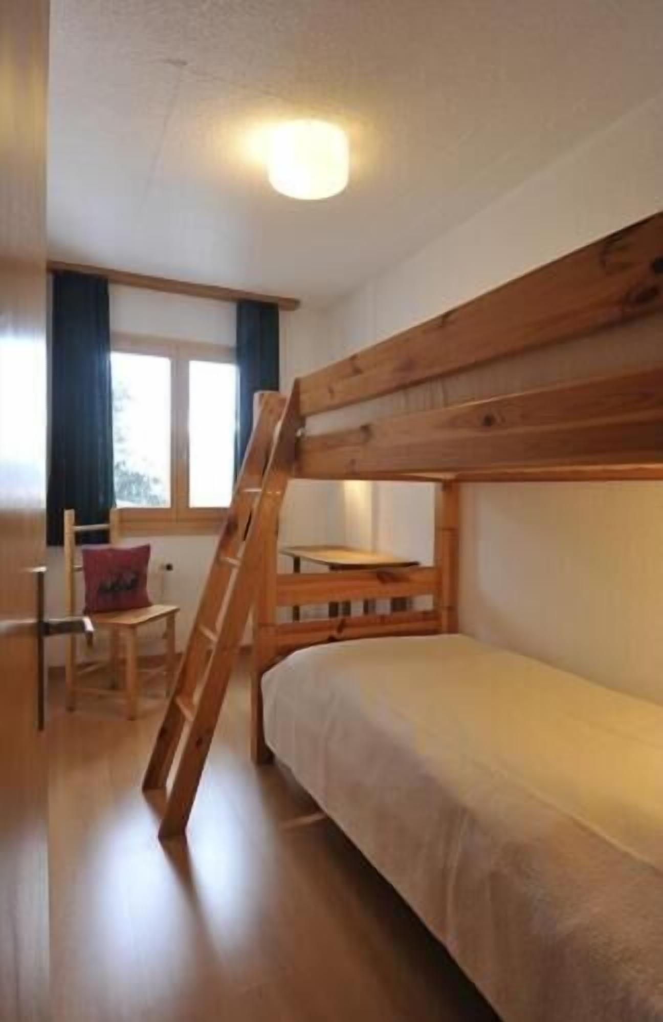 Habitación con literas, una escalera de madera, una silla con cojín y una ventana con cortinas.