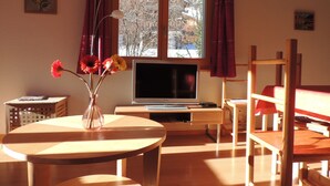 A room with a wooden desk, a television, a window with curtains, and a vase with flowers. - Comfortable 3-room apartment for 4-6 persons : Living room with double sofa-bed - 1 room with 2 beds (Grimentz)