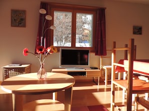 A room with a wooden desk, a television, a window with curtains, and a vase with flowers. - Comfortable 3-room apartment for 4-6 persons : Living room with double sofa-bed - 1 room with 2 beds (Grimentz)