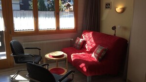 A room with a red sofa, two black chairs, a small round table, and a window with curtains. - Comfortable Studio for 2-4 persons : Living room with sofa-bed (1.40 x 1.90m) and 2 pull-down beds - (Grimentz)