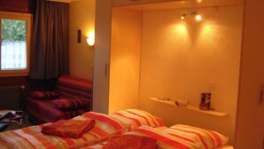 A hotel room with two beds, a window with curtains, a small shelf with items, and a wall-mounted light. - Comfortable Studio for 2-4 persons : Living room with sofa-bed (1.40 x 1.90m) and 2 pull-down beds - (Grimentz)