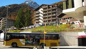 A yellow bus with a CarPostal advertisement parked in front of a building with multiple balconies and a mountain in the background. - Comfortable Studio for 2-4 persons : Living room with sofa-bed (1.40 x 1.90m) and 2 pull-down beds - (Grimentz)