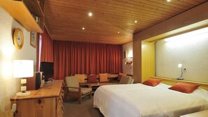 Hotelkamer met een houten plafond, een bed met witte beddengoed en oranje kussens, een bureau met een lamp, een televisie en een raam met nachtlampje.