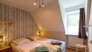 Quarto com teto inclinado, cama com almofadas e toalhas dobradas, mesa de cabeceira com abajur e janela com cortinas.