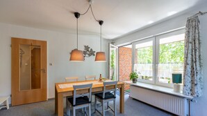 Uma área de refeições com mesa de madeira, cadeiras, luminárias pendentes, um radiador e uma janela com cortinas.