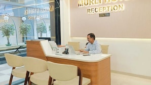 Bancada de recepção no Hotel Murli, com recepcionista, mesa de madeira e parede da recepção com o nome do hotel.