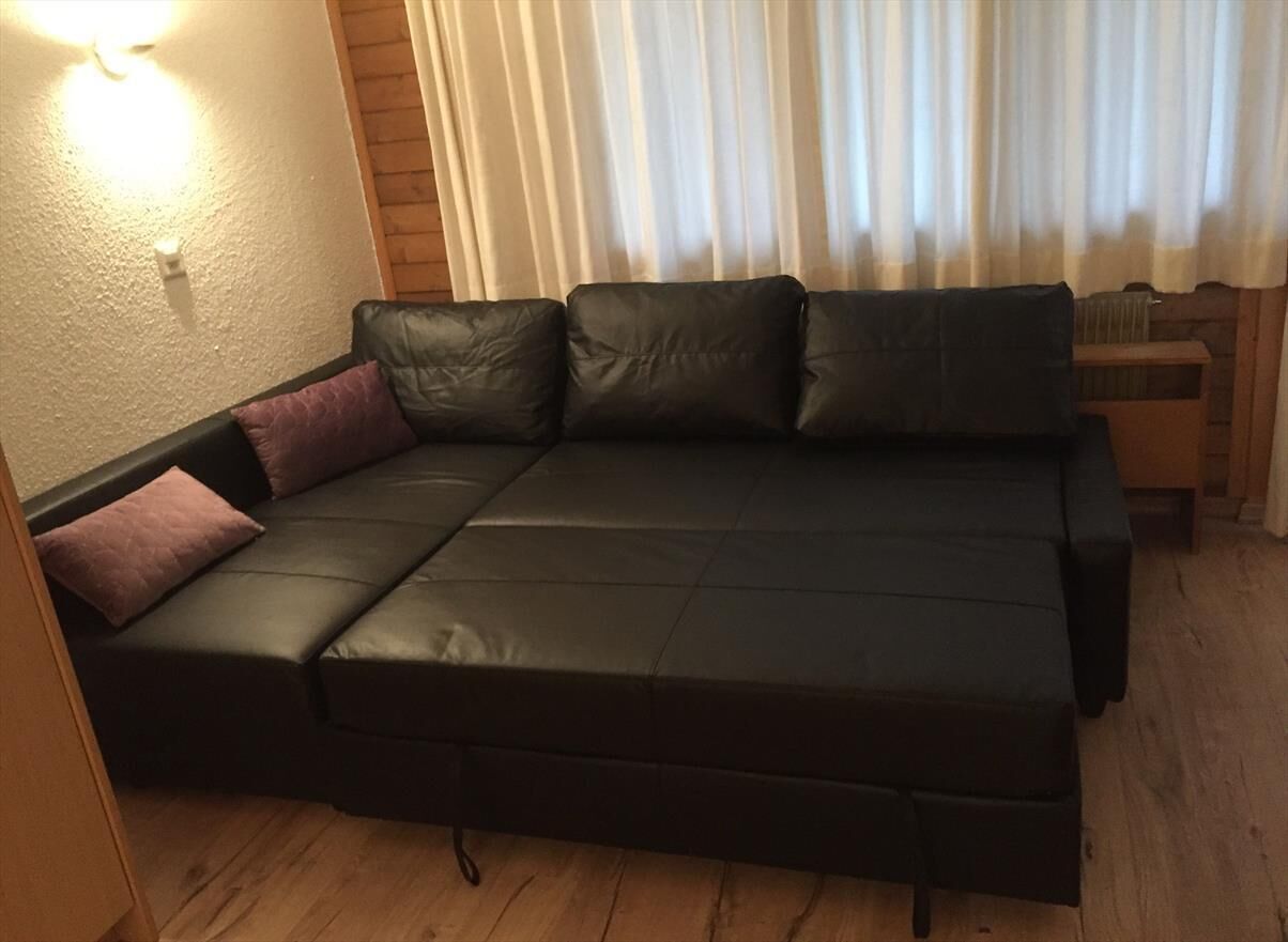 En svart soffa med tre kuddar i ett rum med trägolv, en väggmonterad lampa och ett fönster med gardiner.