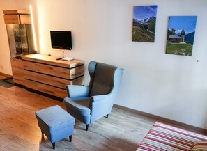 Een moderne kamer met een houten dressoir, een flatscreen televisie, een blauwe fauteuil en een klein voetenbankje. Twee ingelijste foto’s hangen aan de muur.