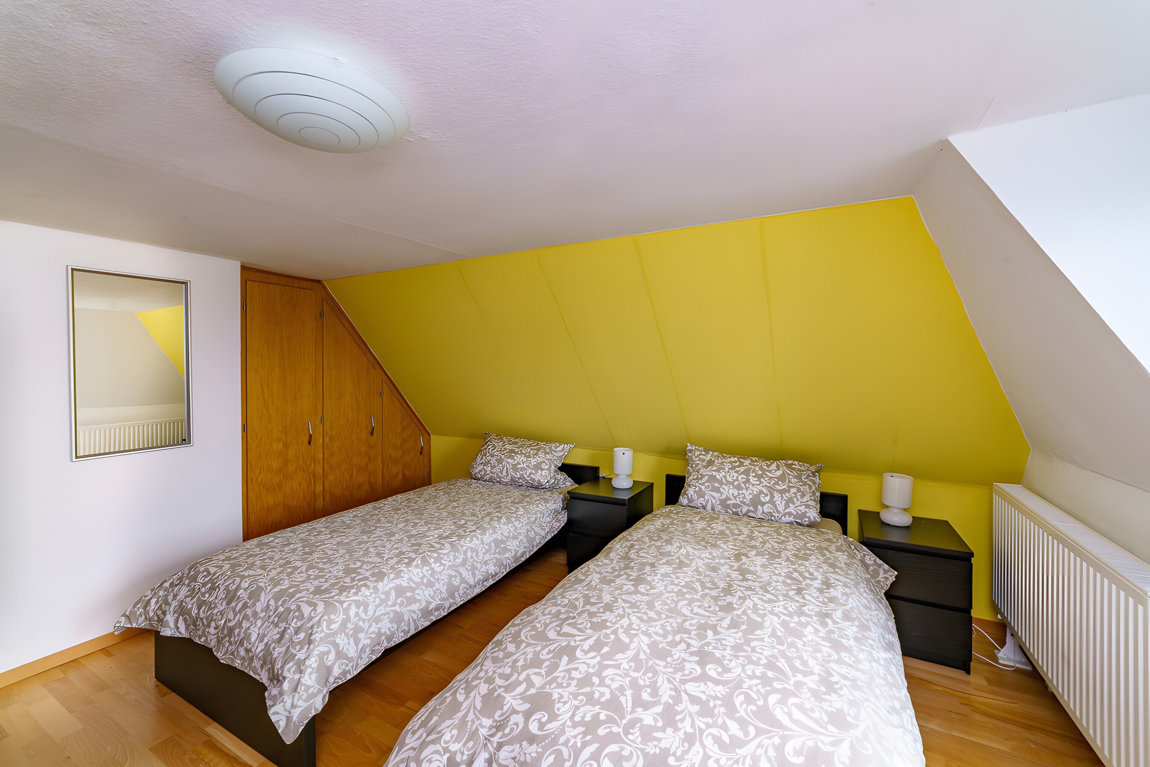 A bedroom with two beds, a wooden wardrobe, a bedside table with a lamp, and a radiator.